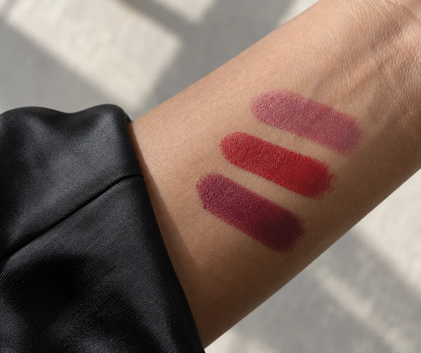 Rose, red and berry lipstick swatches on a medium-brown forearm beside a black satin sleeve