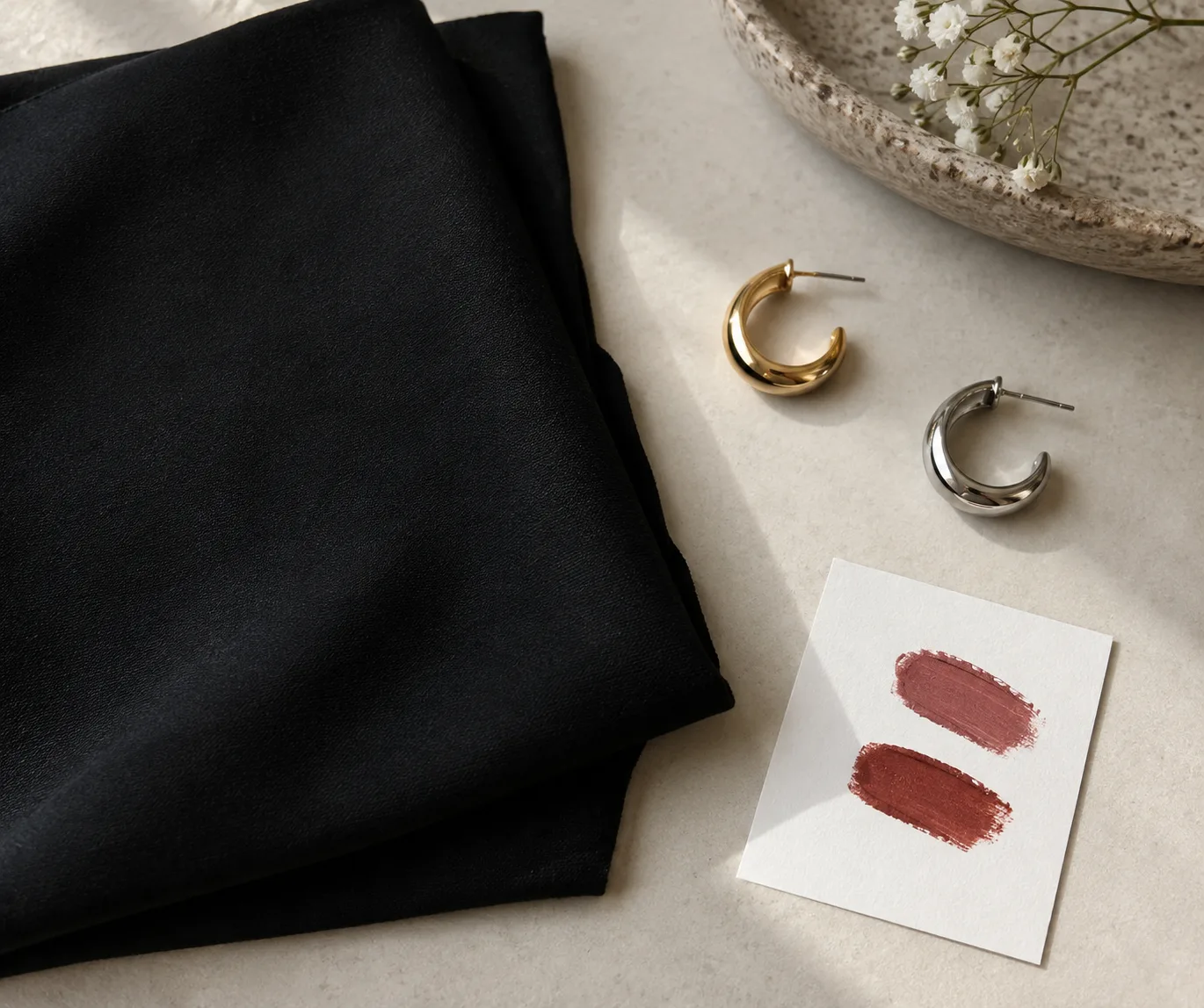 Folded black dress fabric with gold and silver earrings beside two muted lipstick swatches