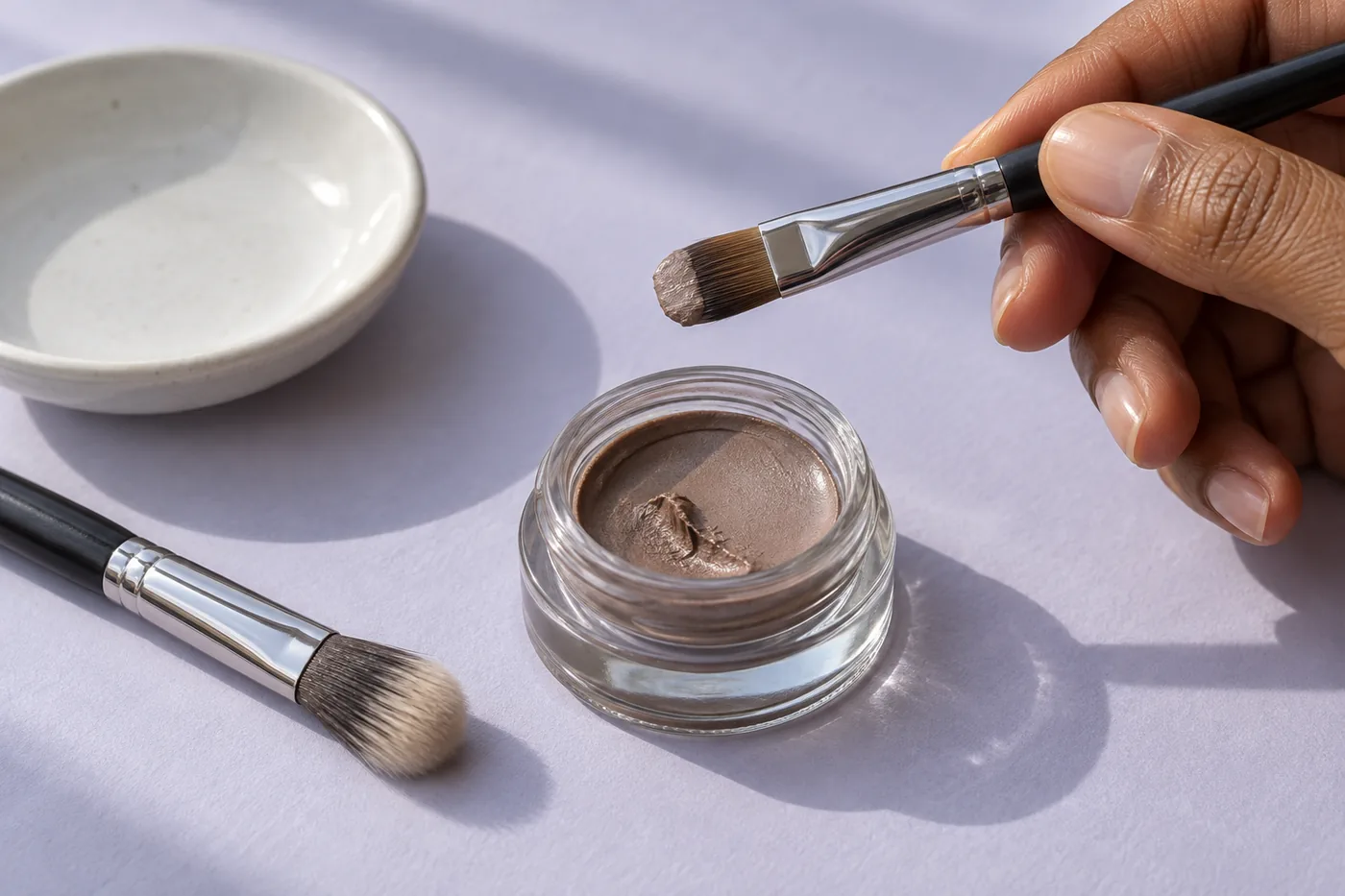 Flat eye brush with visible taupe cream on its tip held above an open unbranded shadow pot