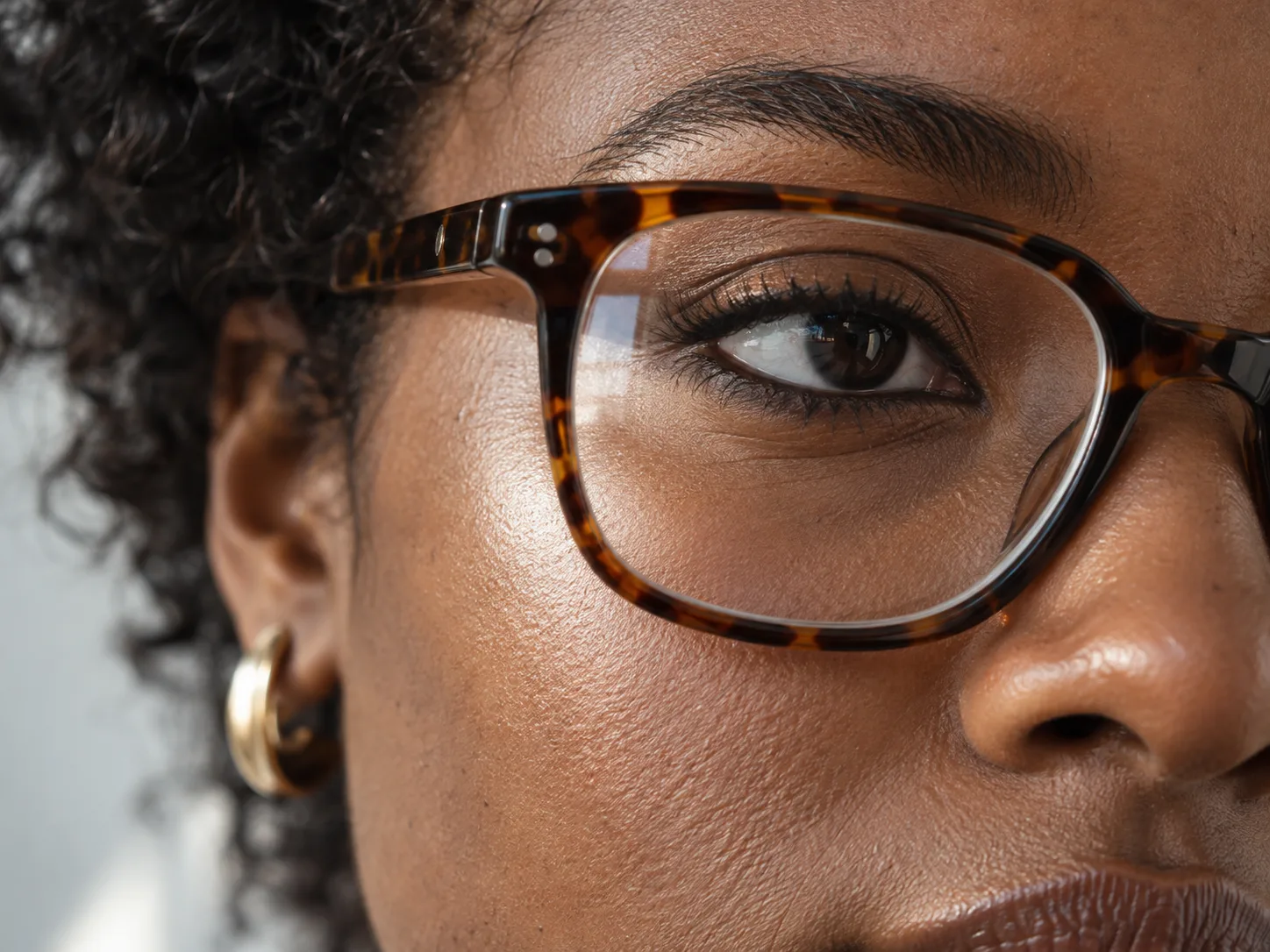 Definierter oberer Wimpernkranz und weicher Bronzeton hinter einer braun gemusterten Brille auf dunkelbrauner Haut