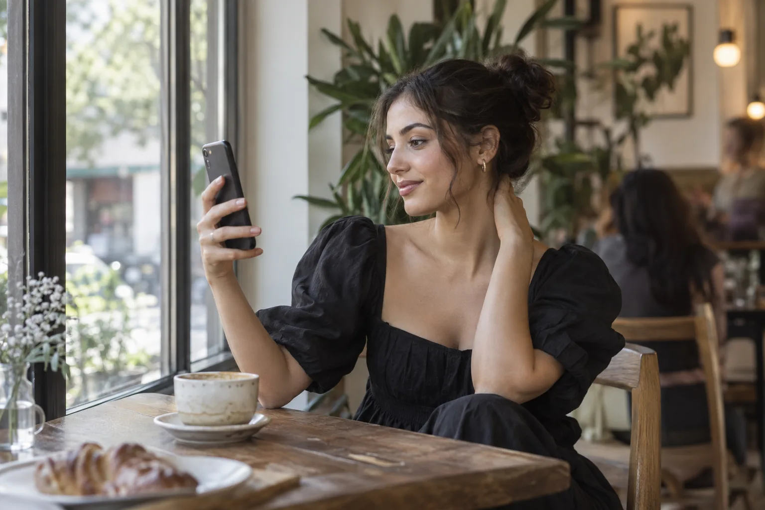 Young woman in a black dress checking her makeup photo on a phone by a bright cafe window