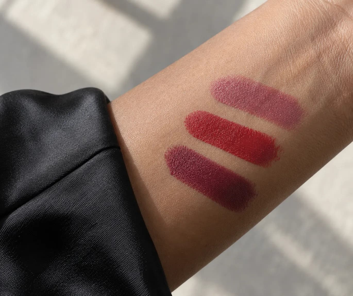 Rose, red and berry lipstick swatches on a medium-brown forearm beside a black satin sleeve