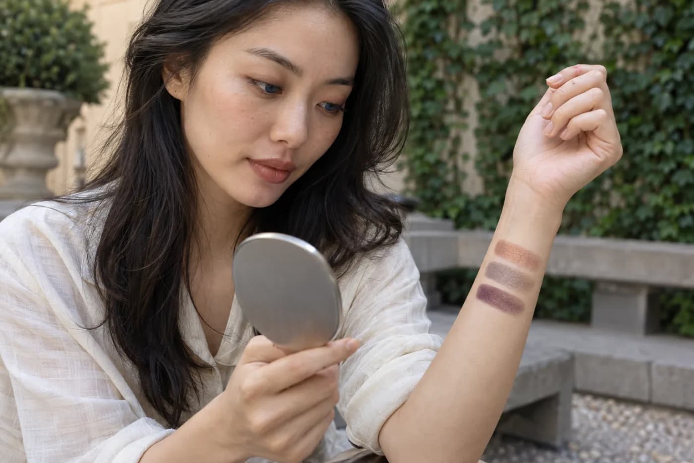 Young woman checking muted copper, neutral taupe and plum eyeshadow swatches on her forearm in open outdoor shade