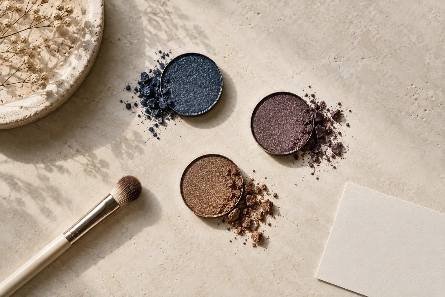 Three unbranded bronze, aubergine and muted navy eyeshadow pans on pale limestone beside a clean blending brush