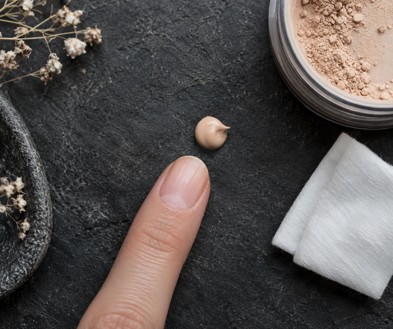 Tiny dot of beige eye base beside a clean fingertip and folded cotton pad on dark stone