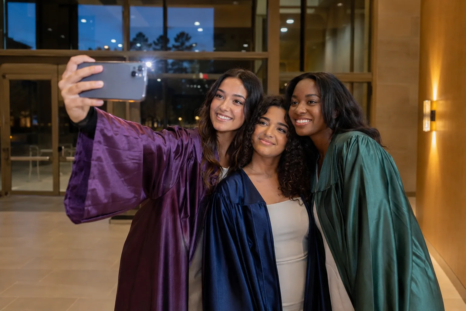 Trois jeunes diplômées en toges prune, bleu marine et verte prennent une photo de groupe au flash dans un hall