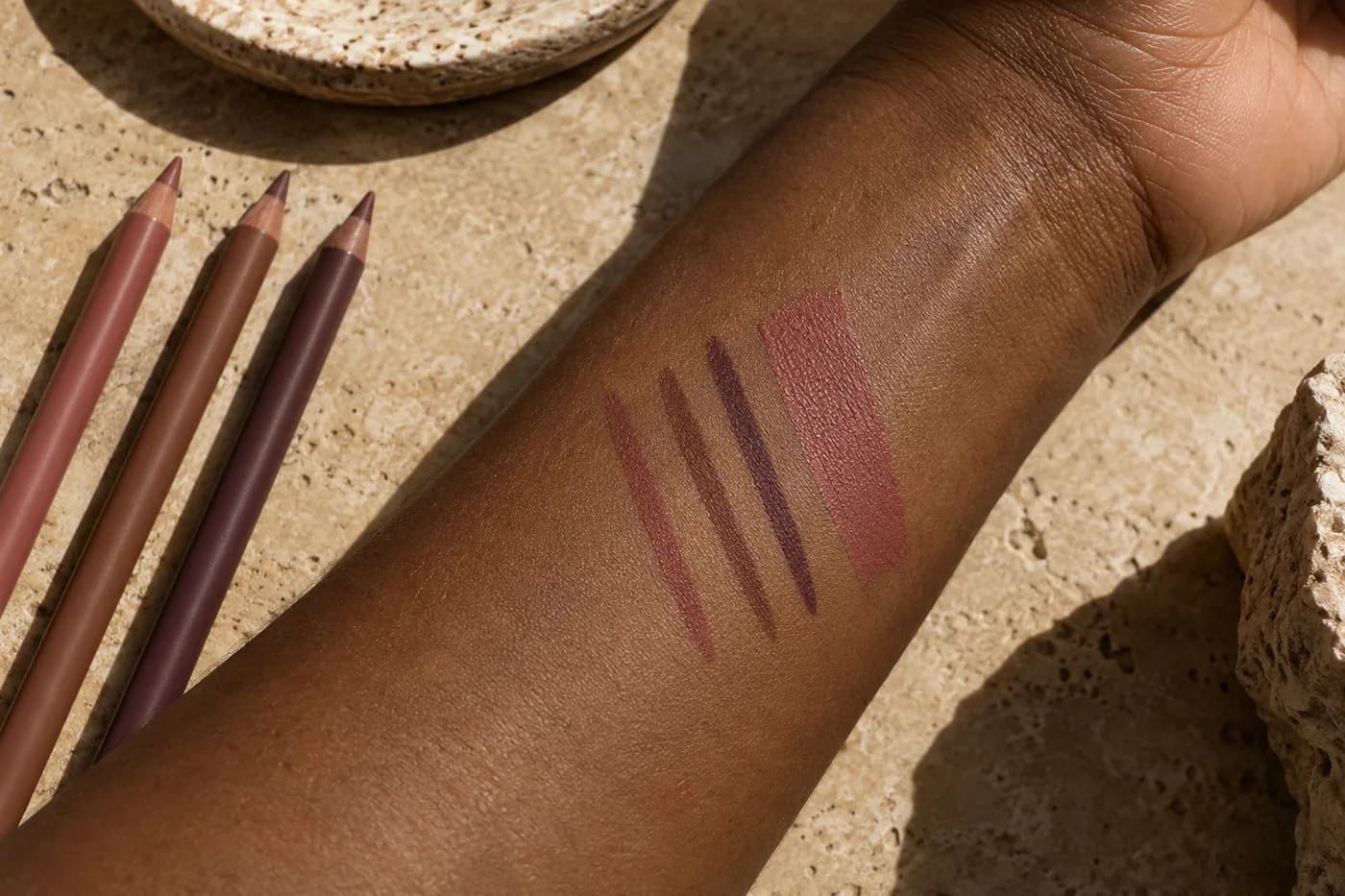 Trois traits fins de crayon et un échantillon de rouge rose poudré sur un avant-bras brun foncé, près de crayons sans marque