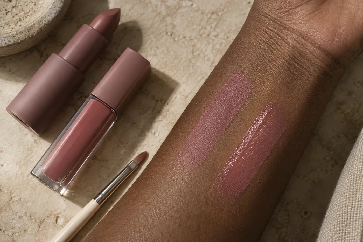 Ähnliche matte und glänzende Roséproben auf einem dunkelbraunen Unterarm neben unbeschrifteten Lippenprodukten und einem Pinsel auf sandfarbener Keramik