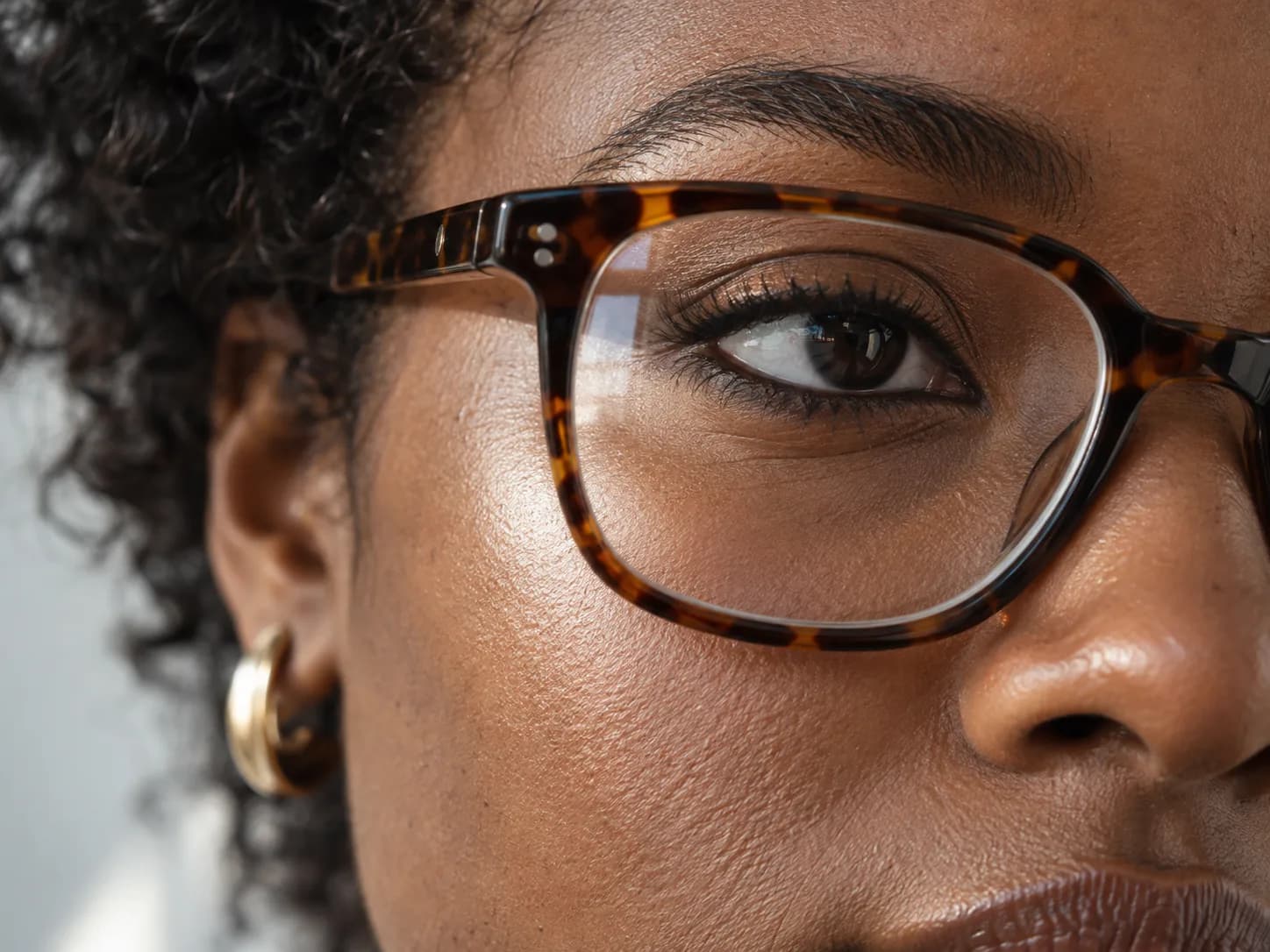 Definierter oberer Wimpernkranz und weicher Bronzeton hinter einer braun gemusterten Brille auf dunkelbrauner Haut
