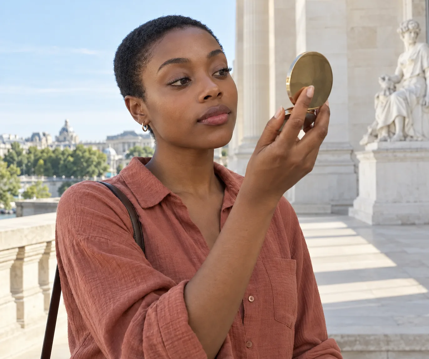 Young Black adult with short hair checks lash definition in a compact mirror on a limestone museum terrace