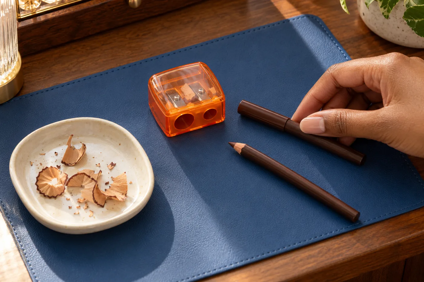 Brown eye pencil, orange cosmetic sharpener, wood shavings and a hand on a blue mat over a walnut table
