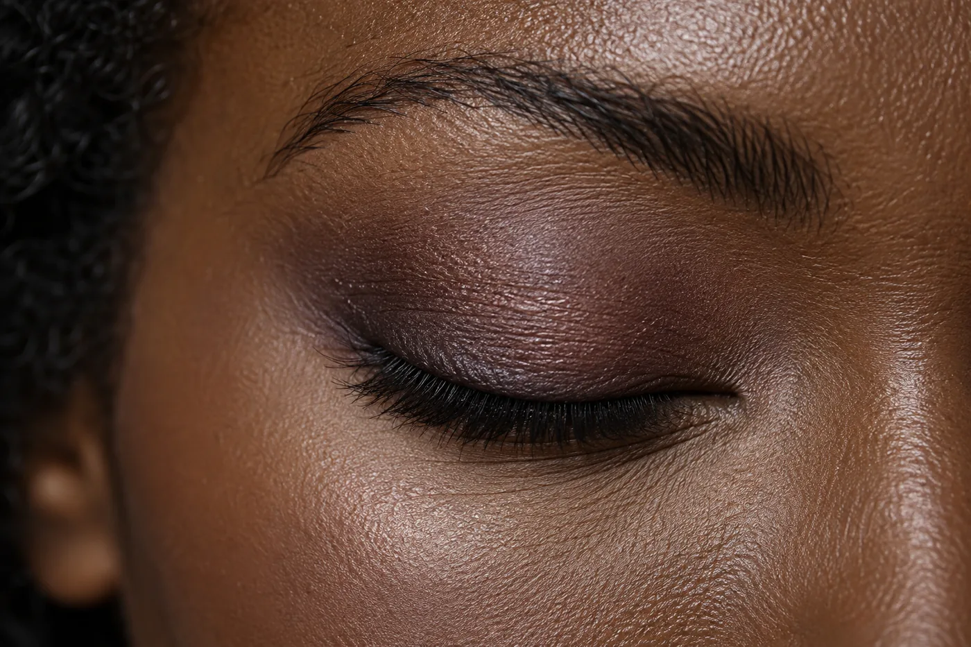 Close view of a softly closed eye with diffused plum-taupe shadow on deep brown skin